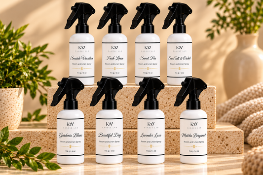 Eight KW Candle Lab Room & Linen Sprays in white bottles with black trigger sprayers, arranged in two rows on beige stone displays. Fragrances include Seaside Vacation, Fresh Linen, Sweet Pea, Sea Salt & Orchid, Gardenia Blanc, Beautiful Day, Lavender Luxe, and Matcha Bergamot.