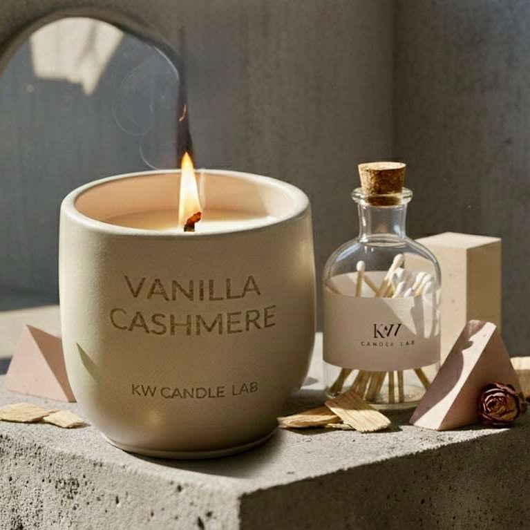 Vanilla Cashmere coconut soy candle with a crackling wood wick displayed on a concrete pedestal alongside a glass match bottle in a warm, modern luxury setting.
