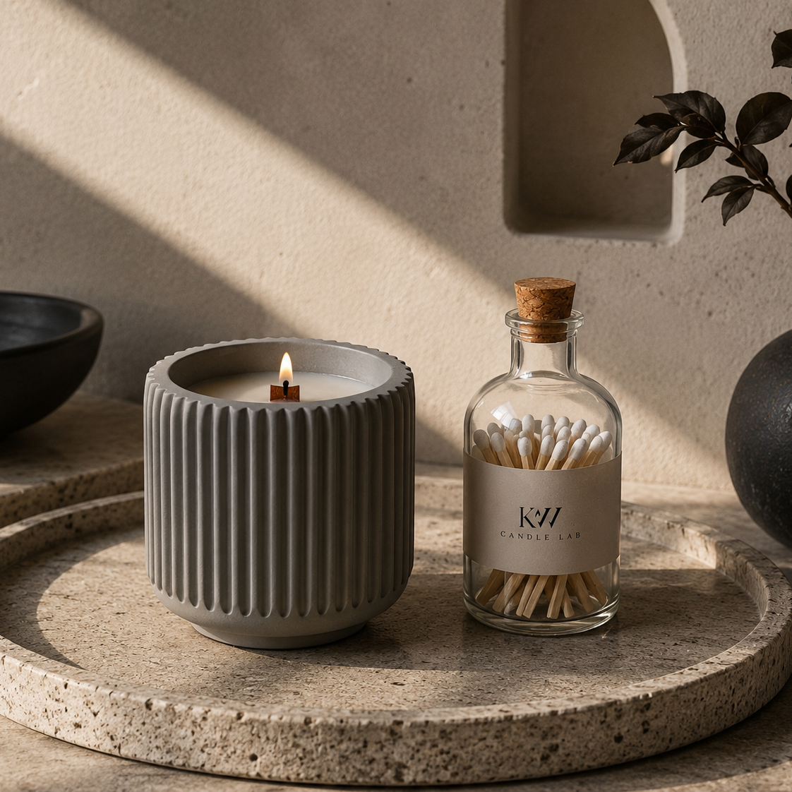 kw candle lab Gray textured obsidian wood concrete candle on a stone surface with a bottle of matches and a plant in the background.